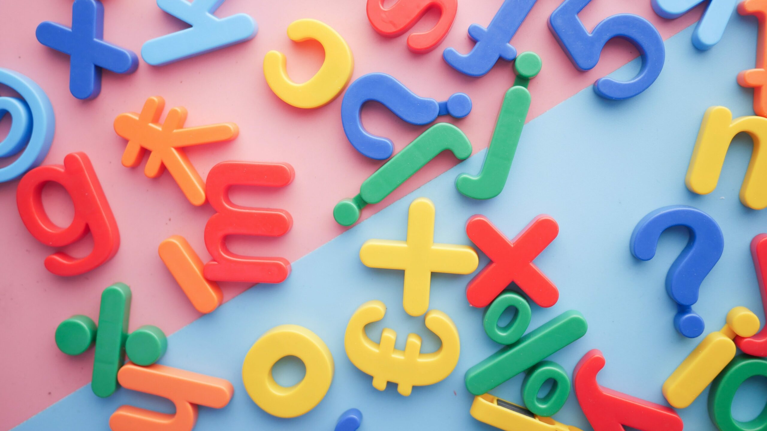 A photo from above of colourful blue, green, yellow, and red letter and symbol magnets spread out on a pink and blue background. 