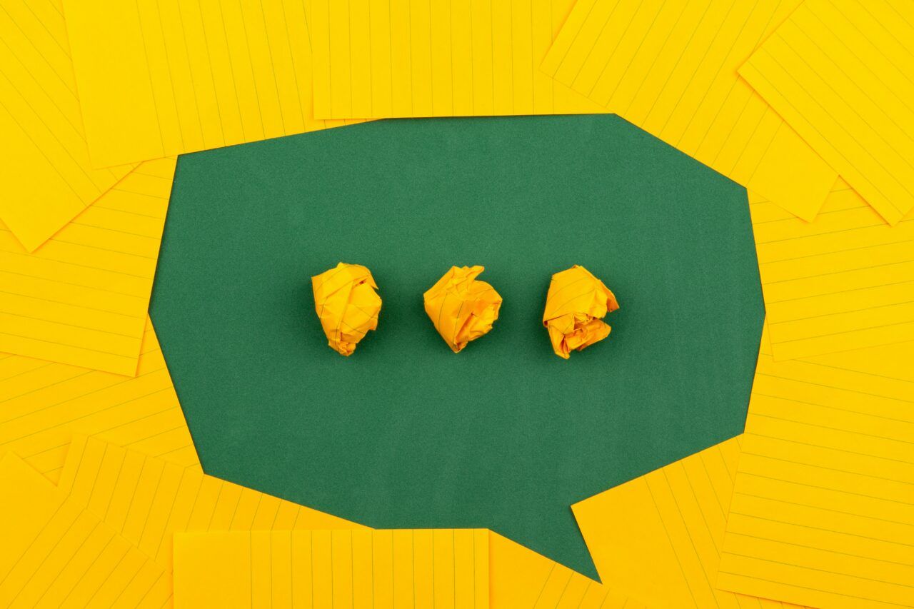 A green speech bubble shaped by numerous yellow Q-cards with three balls of scrunched up yellow Q-cards in the speech bubble to resemble ellipsis.