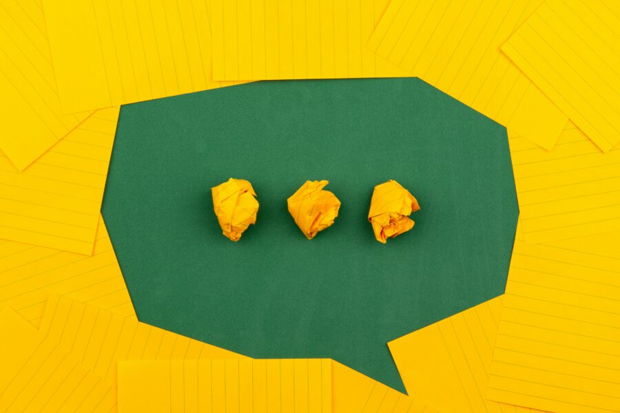 A green speech bubble shaped by numerous yellow Q-cards with three balls of scrunched up yellow Q-cards in the speech bubble to resemble ellipsis.